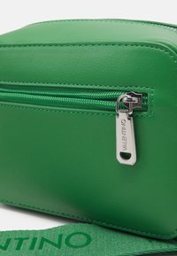 Zelená kožená taška se stříbrným zipem se znakem "VALENTINO" a odpovídajícím zeleným popruhem s embosovaným textem "VALENTINO".