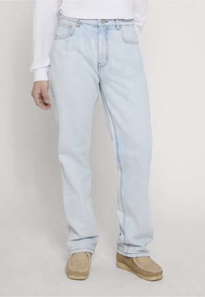 Osoba nosiaca svetlomodré rifle s priamymi nohavicami, béžové mokasíny a biely dlhý rukáv, stojaca pred jednoduchým pozadím.