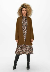 Bruine, knie lange jas met een minimalistisch ontwerp, gedragen over een zwart bloemenjurk, gecombineerd met glanzende zwarte kniehoge laarzen.