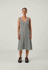 Μέση φόρεμα με ρίγες ασπρόμαυρες, χωρίς μανίκια, με V-λαιμόκοψη, μπροστινά κουμπιά και φουσκωτό φούστα. Φορεμένο με χοντρές μαύρες σαγιονάρες.