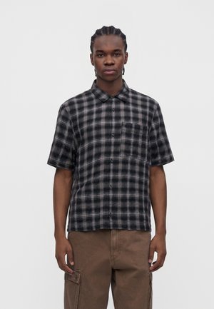 Hombre de pie frente a un fondo liso, vistiendo una camisa de manga corta a cuadros negros y grises y pantalones cargo marrones, mirando al frente con expresión neutral.