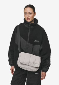 Gray quilted crossbody bag with a flap closure, adjustable strap, and logo detail, worn by a person in a black fleece jacket.