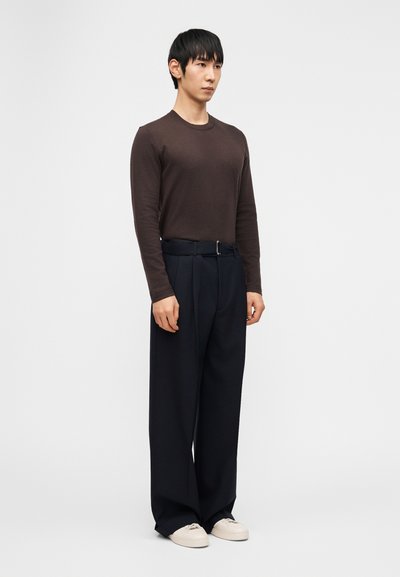 Άνδρας στέκεται φορώντας εφαρμοστό καφέ πουκάμισο με μακριά μανίκια, φαρδιά σκουρομπλε παντελόνια με ζώνη και λευκά αθλητικά παπούτσια, μπροστά από απλό φόντο.