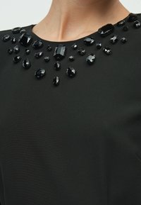 Μαύρο τοπ με στρογγυλό ντεκολτέ διακοσμημένο με διάφορα μαύρα κοσμήματα σε διαφορετικά σχήματα και μεγέθη, τοποθετημένα σε διακοσμητικό μοτίβο.