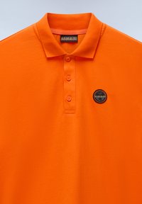 Oranžové polo tričko vyrobené z bavlny s límečkem, dvoubottoným zapínáním a kulatou nášivkou s černým a stříbrným logem na prsou.