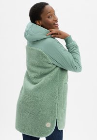 Femme souriante, portant une veste à capuche vert menthe avec des manches matelassées et un dos à texture duveteuse, l égèrement tournée pour montrer les détails du vêtement.