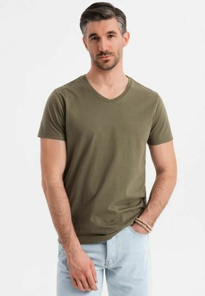 Camiseta de algodón verde oliva con cuello en V y mangas cortas, de corte relajado y textura suave. Llevada con unos jeans azul claro, mostrando un estilo casual.