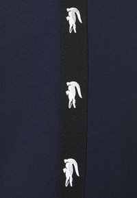 Tissu bleu marine avec une bande verticale noire présentant un motif répété du logo crocodile blanc.