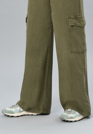 Pantalon cargo ample vert olive avec poches latérales porté avec des baskets vert clair et blanches sur fond gris.