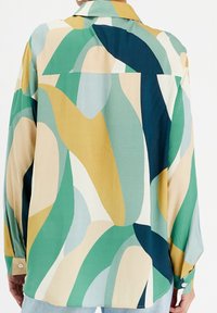 Vue arrière d'une personne portant une chemise à manches longues avec des formes courbes abstraites dans des couleurs vertes, beiges, jaunes et bleu foncé.