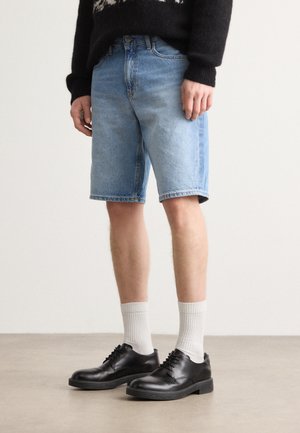 Șorturi din denim de culoare albastru deschis, cu un croi drept, asortate cu pantofi din piele negri și șosete albe cu model în dungi. Fundalul este neutru.