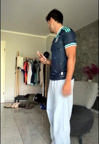 Άνδρας με αθλητική ένδυση κρατάει τηλέφωνο κοντά σε κρεμάστρα με ρούχα και παπούτσια σε ένα σαλόνι με καρέκλα και βάζο με λουλούδια.
