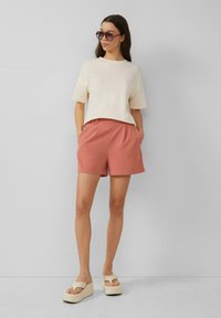 Licht beige katoenen t-shirt gecombineerd met roze op maat gemaakte shorts met zijzakken. Schoeisel bestaat uit sandalen met dikke zolen in roomkleur. Donkere zonnebril gedragen.