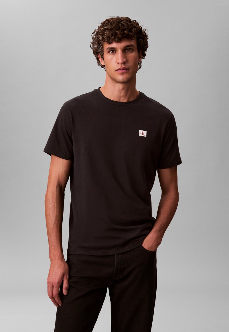 Junger Mann mit lockigem Haar, der ein schwarzes Calvin Klein T-Shirt und schwarze Hosen trägt, steht mit einer Hand in der Tasche vor grauem Hintergrund.