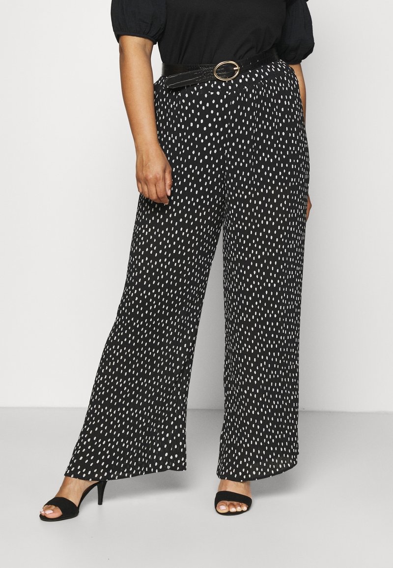 Černé široké kalhoty s bílými puntíky, vyrobené z texturované tkaniny a s opaskem v pase. Spárováno s černými sandály na otevřené špičce.