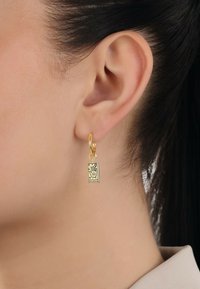 Guldörhänge med en rektangulär charm som har ett tryckt handmönster och texten "VÄRLDEN" längst ner.