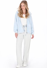 Jasnoniebieska kurtka polarowa z kołnierzem, zapinana na suwak, z opuszczanymi ramionami, zestawiona z jasnoszarymi, wysokim stanem jeansami oraz białymi trampkami.