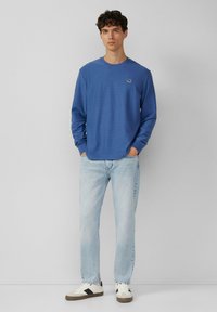 Niebieska koszula z długim rękawem o ryflowanej fakturze z małym logo na klatce piersiowej, zestawiona z jasnoniebieskimi dżinsami i casualowymi białymi trampkami.