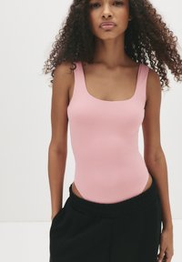 Vrouw met krullend haar in een mouwloze roze bodysuit en zwarte broek, staand tegen een effen witte achtergrond.