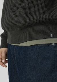 Pull en maille texturée gris foncé avec un ourlet côtelé, superposé à une chemise vert olive, porté avec un jean en denim bleu foncé.