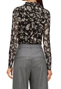Langärmlige Bluse aus durchsichtigem Stoff mit einem grau-schwarzen abstrakten Muster, kombiniert mit hoch taillierten grauen Hosen mit Gesäßtaschen.