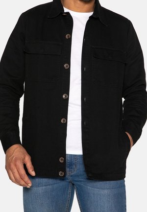 Homme portant une veste noire boutonnée avec poches poitrine sur une chemise blanche et un jean bleu, une main dans la poche, sur fond blanc.