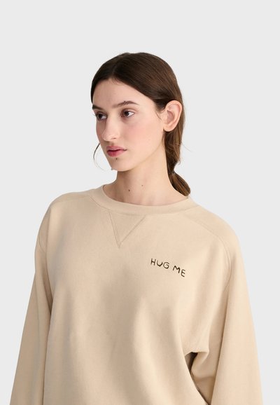 Jeune femme aux cheveux bruns attachés, portant un sweat beige avec les mots "HUG ME" brodés sur la poitrine.