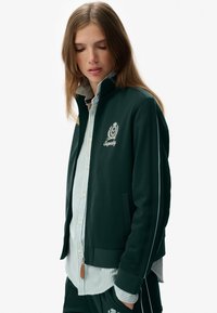 Grøn zip-up jakke med hvide striber, med et logo på brystet, båret over en lyseblå og hvidstribet skjorte. Struktureret stof.