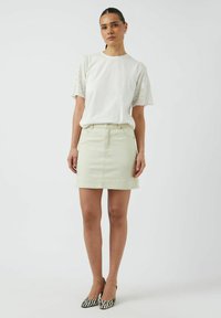 Blusa bianca con maniche in pizzo, abbinata a una gonna di jeans verde chiaro e décolleté a righe bianco e nero, il tutto su uno sfondo neutro.
