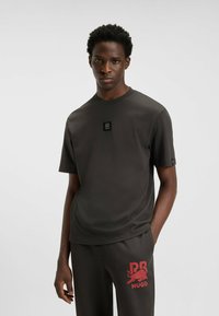 T-shirt a maniche corte di colore grigio scuro con un patch con logo sul petto, collo rotondo e una texture liscia, abbinato a pantaloni scuri con una grafica rossa.