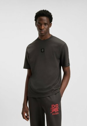 Dunkelgraues kurzärmliges T-Shirt mit einem Logo-Patch auf der Brust, Rundhalsausschnitt und glatter Textur, kombiniert mit dunklen Hosen, die ein rotes Grafikdesign aufweisen.