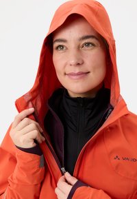 Giacca impermeabile arancione con cappuccio, con interno a fantasia, zip nera e strato interno nero a contrasto, progettata per l'uso all'aperto.