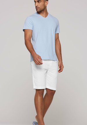 Mann steht mit hellblauem kurzärmligem V-Ausschnitt-Shirt, weißen knielangen Shorts und blauen Slide-Sandalen und schaut nach links.