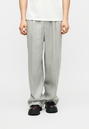 Dolna część ciała osoby w luźnych, jasnoszarych spodniach ze sznurkiem w talii i czarnych masywnych butach, stojącej na białym tle.