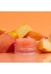 Ferskenfarget leppebalsam i en klar krukke omgitt av ferskenskiver på en oransje bakgrunn. Krukken er merket "NCLA Beauty Balm Babe."