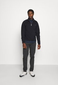 Marine blauwe zip-up sweatshirt over een gestippeld shirt, gecombineerd met grijze broek en sportieve schoenen. De stof lijkt glad met een strak ontwerp.