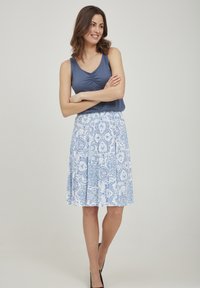 Top sin mangas azul con detalle recogido, combinado con una falda blanca con vuelo que presenta un estampado floral azul. La modelo lleva tacones negros.