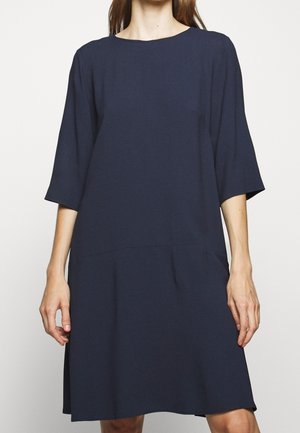 Frau trägt ein locker sitzendes, knielanges dunkelblaues Kleid mit Ellbogenlangen Ärmeln und Rundhalsausschnitt, steht vor einem einfarbigen Hintergrund.