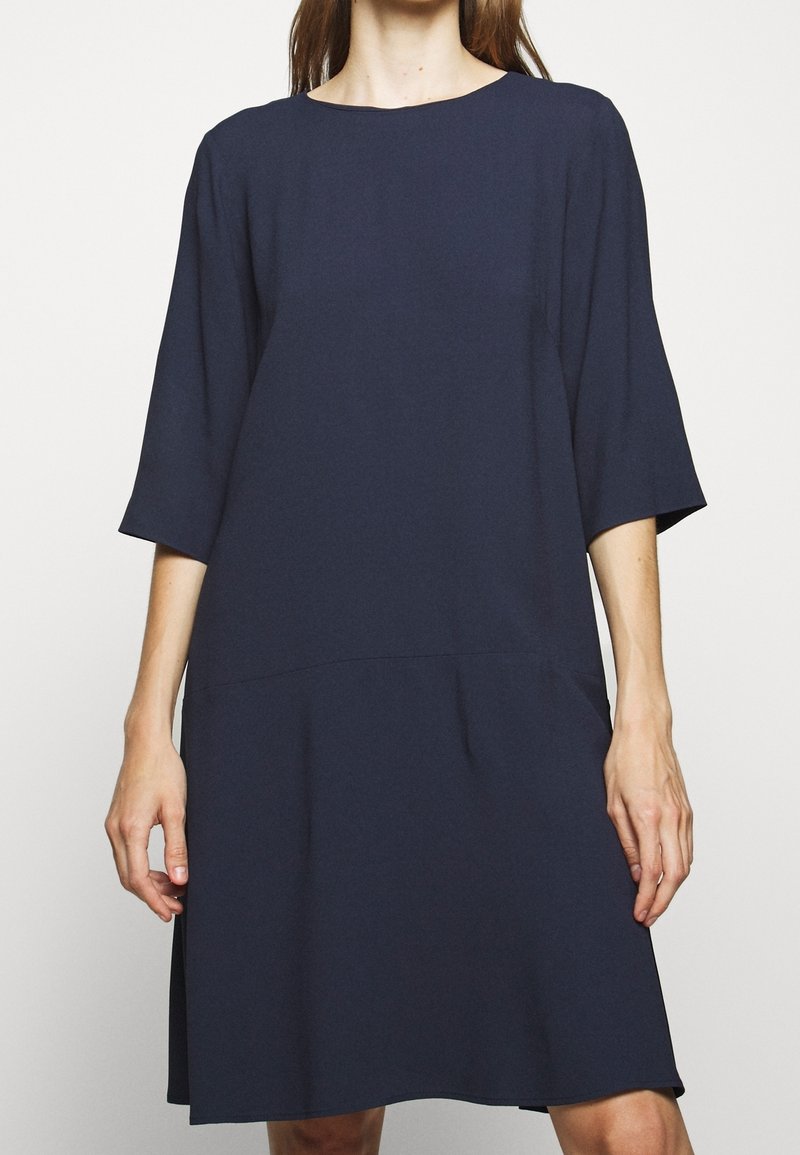 Femme portant une robe bleu marine ample arrivant aux genoux, avec des manches mi-longues et un col rond, debout devant un fond uni.