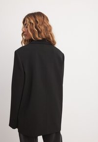 Musta bleiseri, jossa on räätälöity istuvuus, viilennetut kaulukset, yksittäinen nappisuljenta ja kolme nappia hihoissa, joka on varustettu sileällä pinnalla ja suoralla siluetilla.