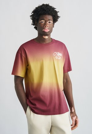 T-shirt a gradiente color bordeaux e giallo, maniche corte, in cotone, con un piccolo logo Nike bianco sul davanti. Il modello indossa pantaloni color crema.