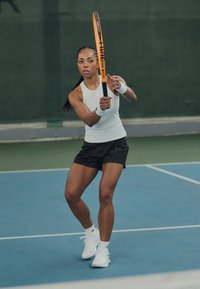 Tennis-pelaaja, jolla on ylleen tiukka valkoinen tankkitoppi ja mustat shortsit, pitää kädessään oranssia ja mustaa mailaa, valmiina lyömään sinisellä kentällä.