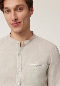 Beige linnen shirt met een Mandarin-kraag, knopen aan de voorkant en een borstzak met geborduurde details. Zachte textuur en lichtgewicht ontwerp.