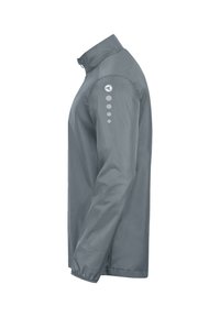 Giacca grigia leggera con colletto alto, polsini elasticizzati e una texture liscia. Presenta quattro elementi circolari sulle spalle.