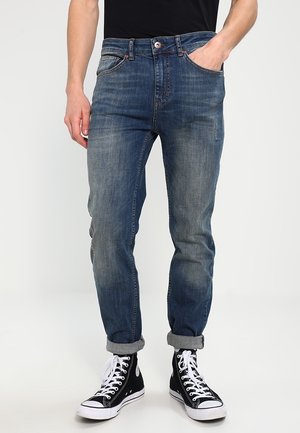 Mann trägt blaue Jeans mit Umschlag, Hand in der Tasche und schwarze High-Top-Sneaker mit weißen Schnürsenkeln vor einfachem Hintergrund.