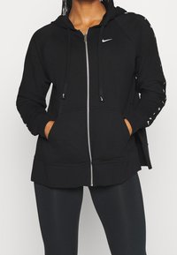 Personne portant un sweat à capuche zippé noir Nike avec logo blanc, les mains dans les poches avant, associée à un legging noir.