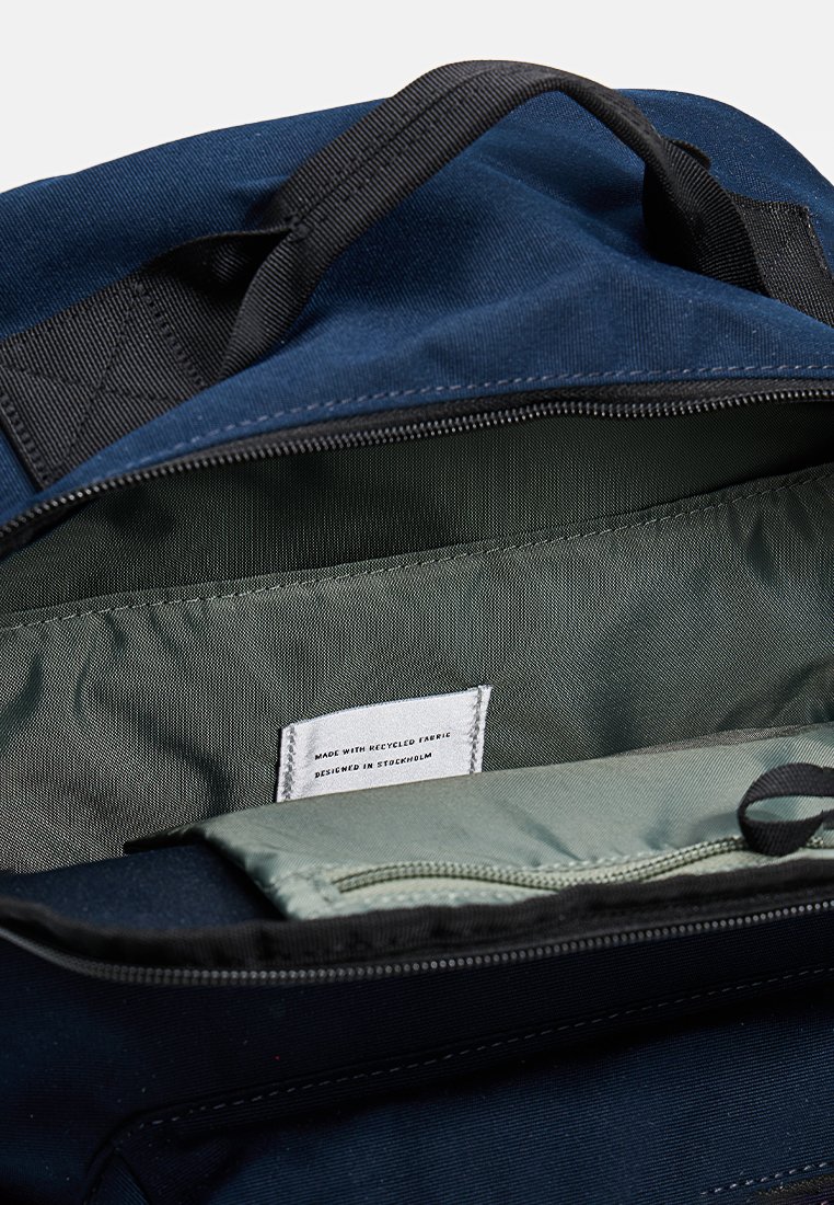 Bolso de tela azul marino con apertura con cremallera. El interior cuenta con un forro suave de color verde oscuro y una etiqueta que indica el uso de material reciclado.