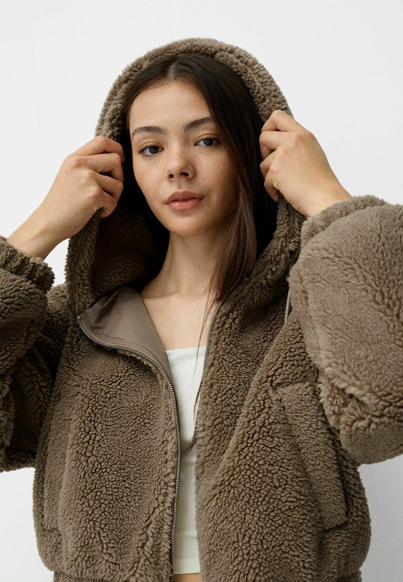 Veste zippée en polaire marron avec capuche, dotée d'une texture douce et d'une coupe oversize. L'intérieur est en matériau beige lisse.