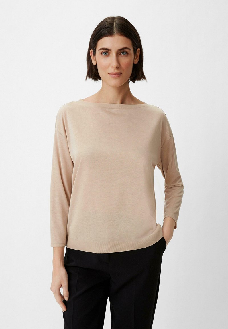Haut en maille beige avec encolure bateau et manches trois-quarts, présentant une texture lisse et une coupe décontractée, associé à un pantalon noir.
