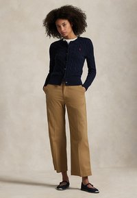 Kvinde med krøllet hår iført en navy kabelstrikket cardigan, hvid skjorte, tan vidde bukser og navy espadrille-flade sko, stående med hænderne i lommerne.